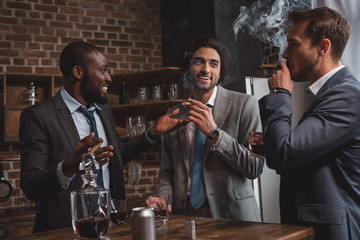 smiling multiethnic male friends in suits talking, drinking whiskey and smoking cigars