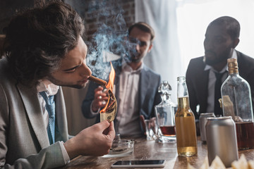 young businessman holding dollar banknote and smoking cigar while partying with friends