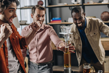 excited multiethnic men building tower from bottles and glasses while having fun together at party
