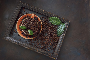 Close-up photo of cup with aroma coffee beans and fresh green leaves in frame on black table background