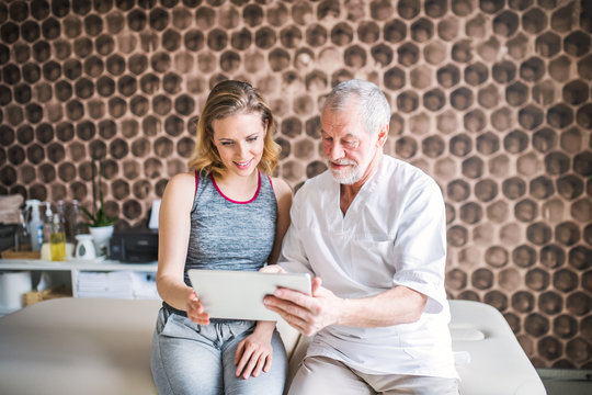 Senior Male Physiotherapist And Young Woman With Tablet Sitting.