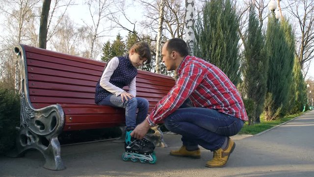 father helping daughter dresses roller skates on bench in park. leisure time. parental care. family relations and support