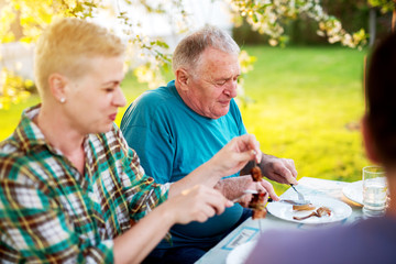 Elderly man and mature woman are sitting at the table outside under the tree and eating .