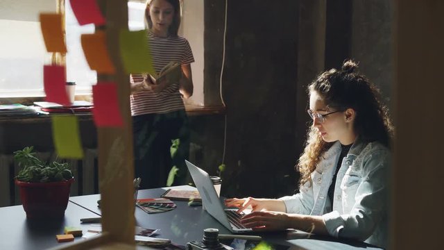 Two attractive girls are working together in modern loft office. Curly businesswoman is typing on laptop sitting at table while her colleague is reading book standing near window