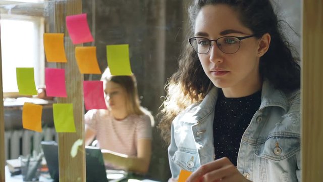 Close-up shot of attractive young businesswoman standing and sticking colourful memos on glass board. Her colleague is sitting and working in background. Creative work concept.