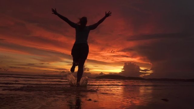 Slow motion, silhouette, low angle cheerful woman runs into the ocean and towards the lovely sunset with her arms outstretched