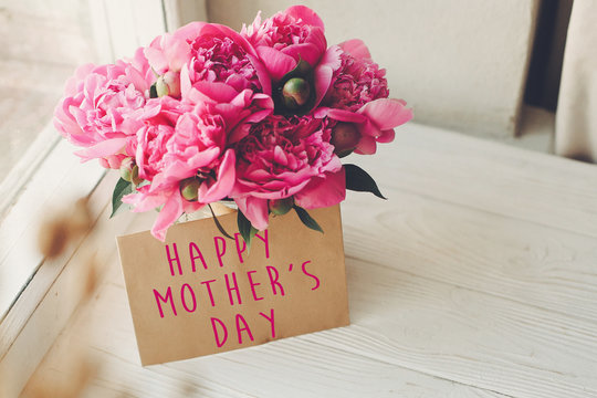 happy mother's day text on craft card and pink peonies bouquet on rustic white wooden window in light. floral greeting card concept. mothers day. soft tender spring image