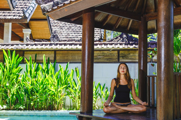 Female meditating by the pool, Bali, Indonesia.The girl sits in a gazebo at a bali in a lotus pose and meditates with a view of the pool.