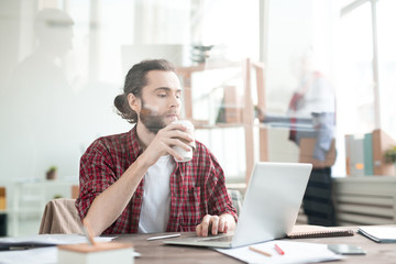 Casual office worker typing on laptop and drinking coffee at the table