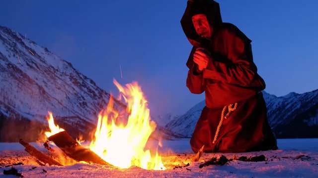 Alone monk in brown cloaks of the order of St. Francis is warming hands by the fire and prays