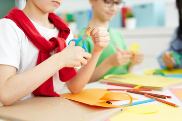 Kids sitting at school desk and cutting colorful paper with scissors while making origami on art...