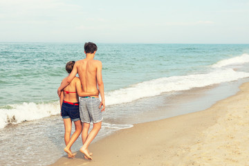 Two teenagers: a girl and a boy with blond hair, dressed in a swimsuit are walking on a sea beach. Back view.