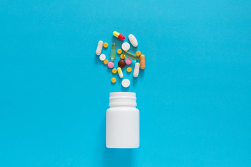 Colorful different tablets spilled from a white plastic bottle on an empty blue back ground