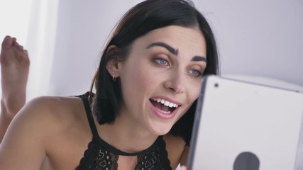 Young sexy caucasian brunette girl in black underwear using a tablet, lies on stomach, sexy feet in the background, white bedroom background 50 fps