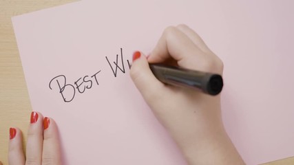 Female hands writing best wishes in capital letter on pink paper with a black marker