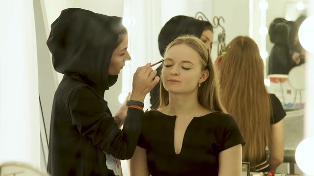 Woman visagiste doing makeup eyebrows on face young model in beauty studio. Woman visagiste coloring eyebrows on face makeup model front mirror in dressing room.