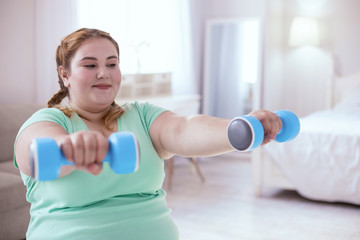 Dumbbells exercises. Plump young woman smiling while holding dumbbells opposite her