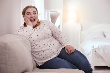 Amazing news. Surprised young woman sitting on the couch while hearing excellent news