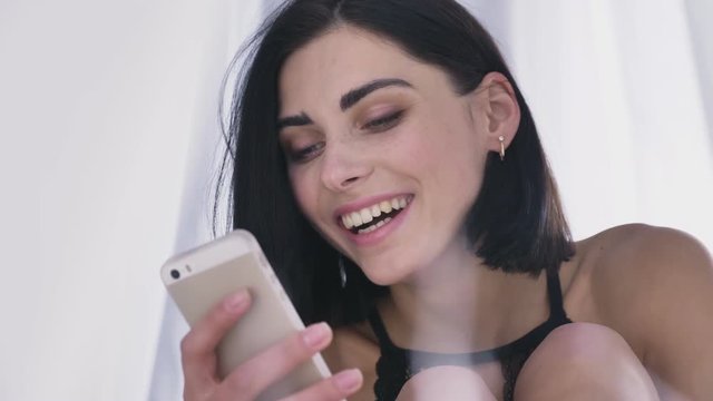Portrait of young sexy caucasian brunette girl in lingerie using smartphone reading, smiling, laughing, holds head with hand, white background 50 fps