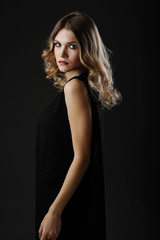 Fototapeta na wymiar Beautiful blonde hair woman in black dress looking back while posing in the studio on black background. Fashion portrait