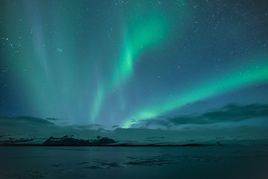 Northern lights aka Aurora Borealis glowing on sky in Jokulsarlon glacier lagoon with snow capped mountains in background at night in Iceland