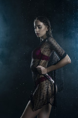 Beautiful wet slim girl wearing a red lingerie and black translucent veil posing sideways in rain water drops in a studio on black background in a theatrical smoke