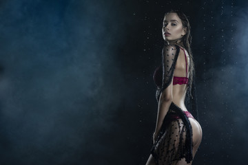 Fototapeta na wymiar Young wet slim girl wearing a red lingerie and black translucent veil posing sideways in rain water drops in a studio shows her beautiful butt on black in a theatrical smoke