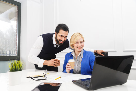 Two business colleagues work with laptop holding cups of coffee in white office interior
