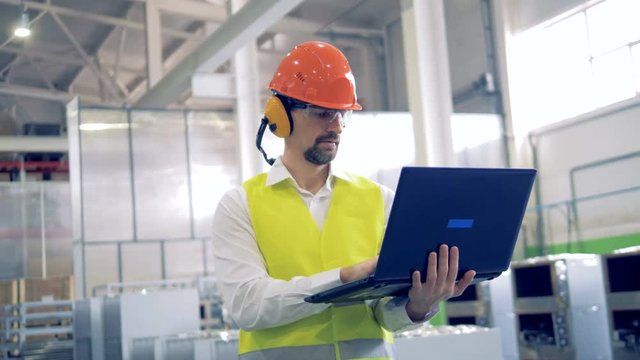 A man in hard hat works on his laptop at a industrial factory.