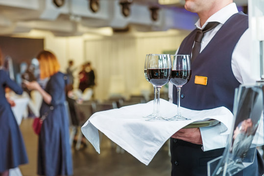 Professional waiter in uniform holding a tray with glasses of vine at business event. Catering or celebration concept. Service at corporate meeting, party, weddings. Selective focus, space for text.