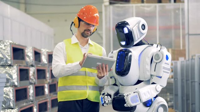 Male worker talks to a robot, showing it information on a tablet, then types on a robot's monitor. 4K.