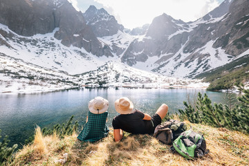 Fototapeta na wymiar Father and son backpackers sit near the mountain lake and enjoy mountain snowy peaks