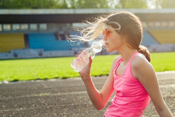Beautiful teenage girl resting after workout at stadium, drinking water