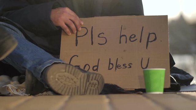 Close-up of beggar male's legs and hands holding cardboard sign asking for help. Homeless man sitting on street begging for money while people's legs walking and passing by.