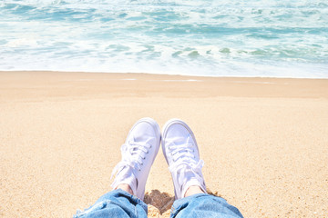 Naklejka premium Female feet in jeans and white sneakers on beach. Walk along the beach