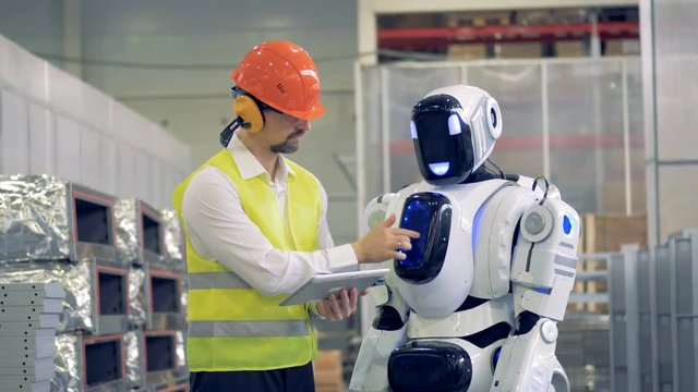A worker shows a cyborg some gestures to copy, then they give each other a hi-five. 4K.