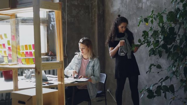 Female friends and business partners are working together in modern office. Blonde is sitting at table and collecting pictures, brunette is watering plants.