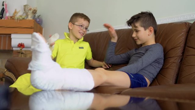 Two brothers with broken leg and hand sitting on sofa at home and talking
