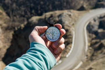 Close-up A man's hand holds a pocket compass against the backdrop of a mountain road and forest. The concept of outdoor navigation © yanik88