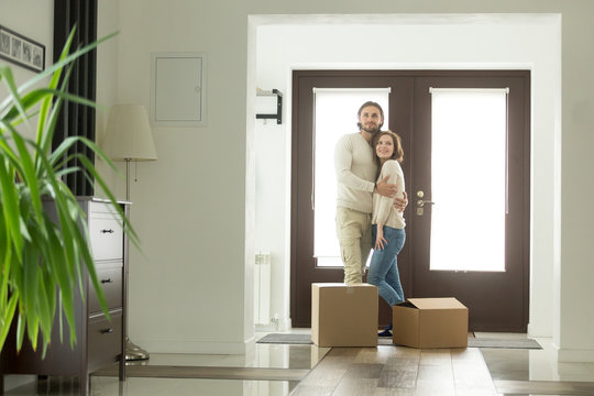 Moving day concept, happy young couple owners hugging embracing in new home standing with boxes in modern hallway of own big luxury house, mortgage loan investment, buying real estate and relocation