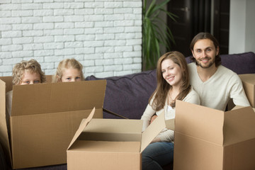 Portrait of happy family with kids on moving day, cute children son and daughter playing peeking from boxes, parents looking at camera sitting on sofa together, relocation packing unpacking concept