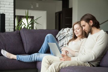 Obraz premium Young family couple using laptop discussing news or doing online shopping sitting together on sofa at home on weekend together, serious man and woman browsing web searching for new sale offers