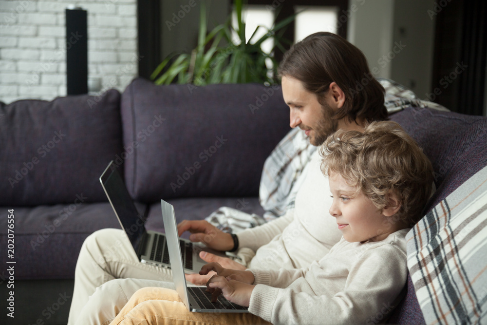 Wall mural curious smiling child typing on laptop looking at screen with dad on sofa, daddy and little boy usin