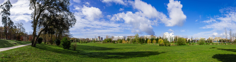 Fototapeta na wymiar Panorama of a large empty green grass lawn field, with a view of the city in Parque da Devesa Urban Park. Blue sky with large white clouds. Vila Nova de Famalicao, Portugal