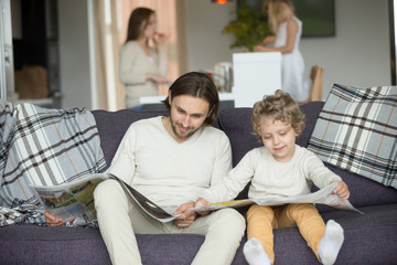 Little son pretending reading newspaper sitting on couch with father, cute funny kid imitating dad to be alike spending weekend with family, child boy learning to read discussing news with daddy