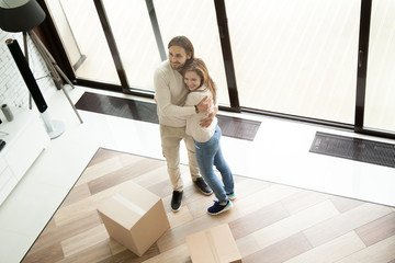 Fototapeta premium Young happy couple embracing arriving in house on moving day, smiling excited family hugging standing in new home with boxes, buying real estate, mortgage and relocation concept, top overhead view