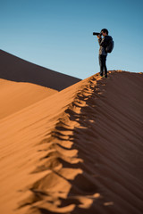 Young male traveler and photographer standing on the top of sand dune photographing sunrise or...
