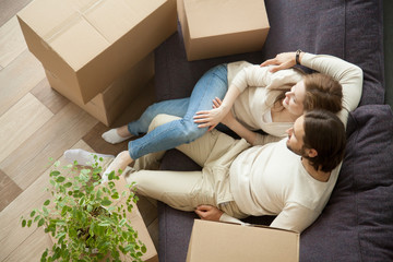 Relaxed couple resting on sofa on moving day, young renters relaxing on couch moved into apartment with boxes, happy satisfied home owners enjoying first day in new house after relocation, top view