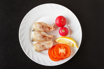 Steak fish with red radish, slice lemon and chopped tomatoes on white plate. Black background. Top view