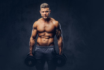 Obraz na płótnie Canvas A handsome shirtless tattooed bodybuilder with stylish haircut and beard, wearing sports shorts, posing in a studio. Isolated on a dark background